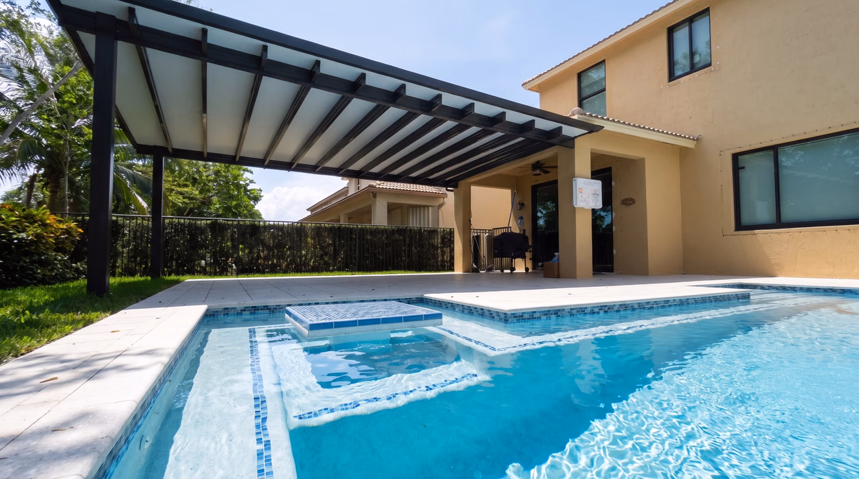 Slatted aluminum pergola shading the lanai beside a pool with a raised glass mosaic spa in a South Florida backyard.
